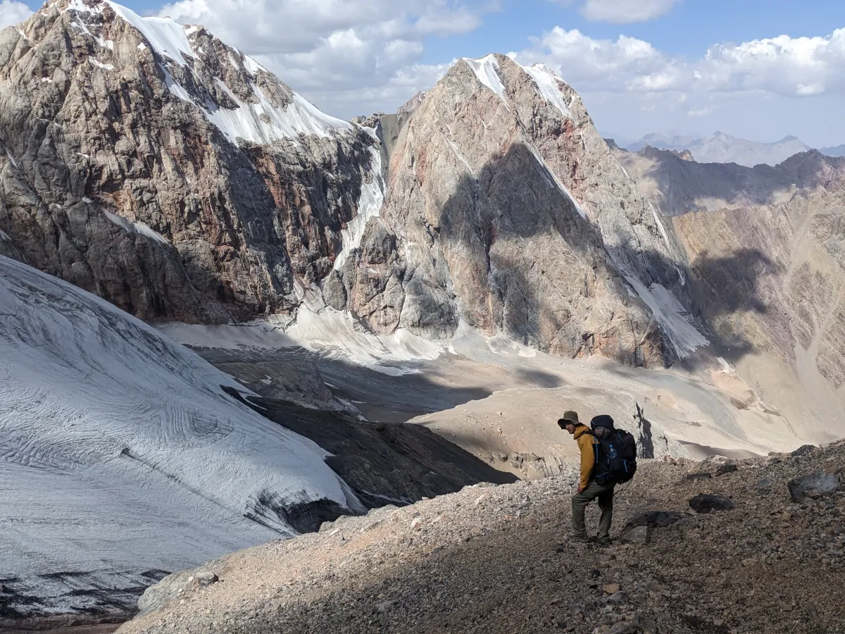 Hiker with yellow jacket standing on a rocky slope on a backdrop of rough barren mountain peaks with a patchy lighting pattern
