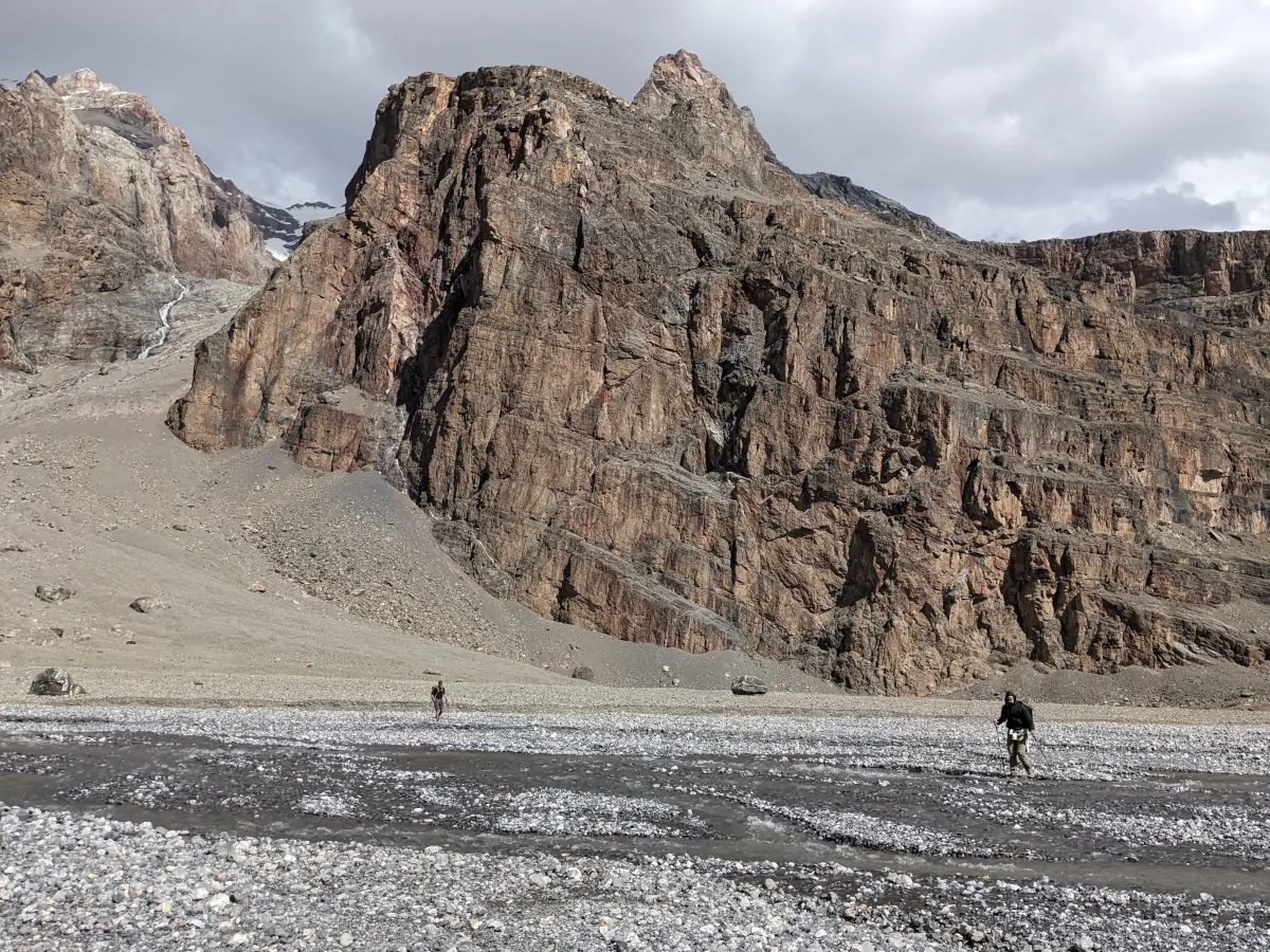Two hikers on the rocky bed of a braiding river with a vertical rock cliff in the background