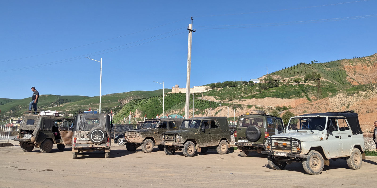Seven UAZ green jeeps parked with the Baljuvon fortress perched on a hill in the background