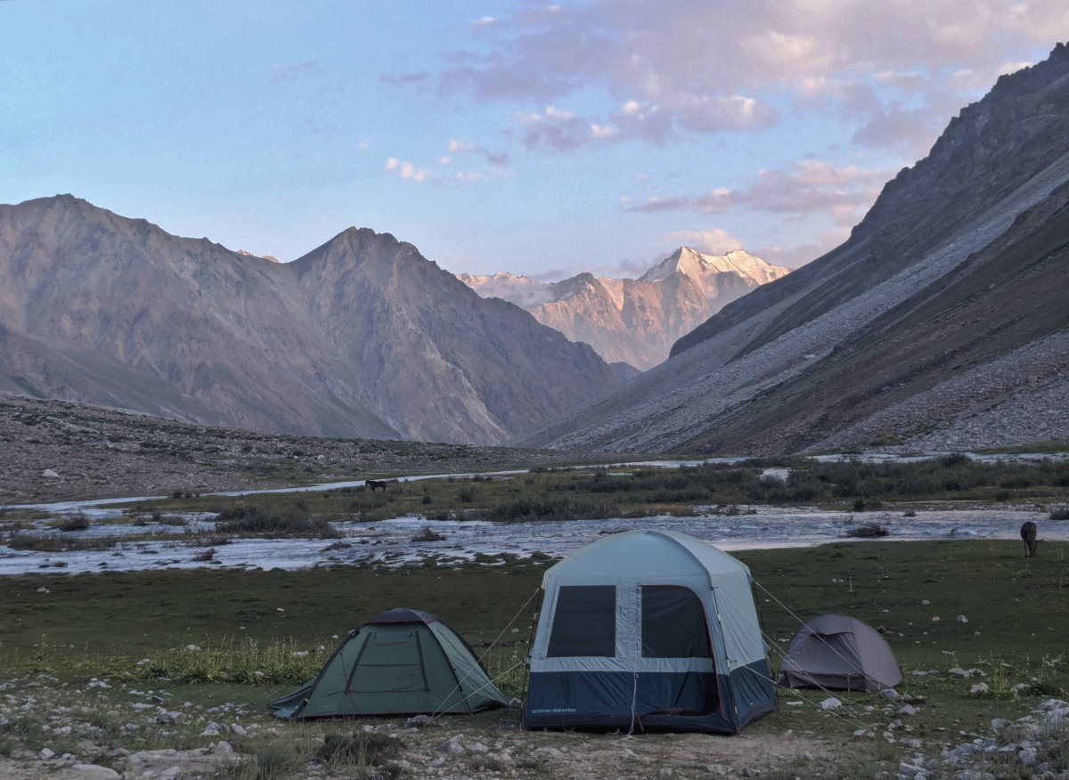 Three tents pitched in the Andaravadzh Valley in the Pamirs in front of a winding river with mountains in the background that are highlighted by a setting sun